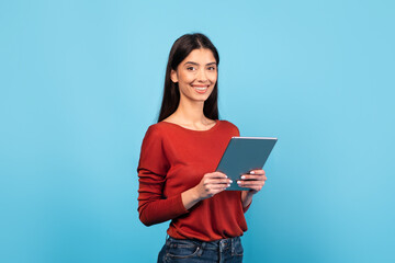 Fototapeta premium A millennial woman with long brunette hair wears a red sweater and smiles while holding a tablet. The bright blue background enhances her cheerful demeanor, creating a lively atmosphere.