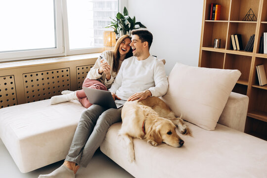 Couple laughing using laptop with golden retriever dog on sofa at home