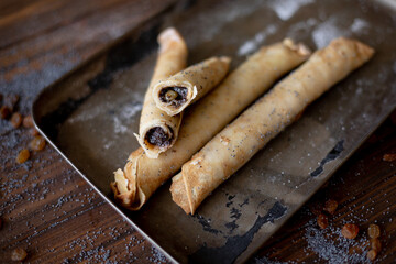 Golden-brown rolled pancakes with a rich poppy seed and raisin filling displayed on a vintage metal tray. The wooden surface is dusted with poppy seeds and raisins for a rustic, appetizing presentatio