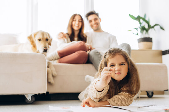 Little girl drawing with concentrated expression and parents relaxing on sofa with dog - Powered by Adobe