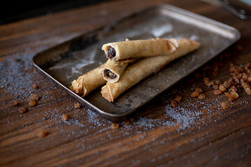 Rolled pancakes with sweet prune filling and poppy seed topping, served on a rustic baking tray, surrounded by raisins and powdered sugar on a wooden table.