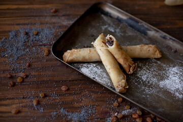 Rolled pancakes with sweet prune filling and poppy seed topping, served on a rustic baking tray, surrounded by raisins and powdered sugar on a wooden table.