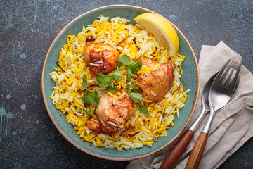 Delicious indian chicken biryani with chicken, garnished with fresh cilantro and lemon, in a bowl on a rustic table, accompanied by cutlery. Authentic dish of India