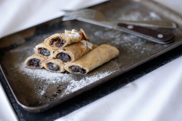 A neat stack of thin pancakes filled with dark poppy seed filling, garnished with powdered sugar  on a dark metal tray. The white tablecloth and cutlery create an elegant dining setup.