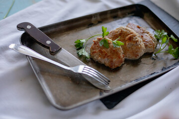 A trio of juicy meat patties is artfully arranged on a vintage baking tray, garnished with fresh parsley leaves. Fork and knife are placed next to the tray on a rustic white cloth, highlighting the ho