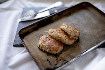 Freshly fried meat patties rest on a slightly worn metal tray, with visible oil drops enhancing their golden appearance. The cutlery peeks from the background on a dark surface, giving a rustic, homel