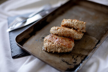 Freshly fried meat patties rest on a slightly worn metal tray, with visible oil drops enhancing their golden appearance. The cutlery peeks from the background on a dark surface, giving a rustic, homel