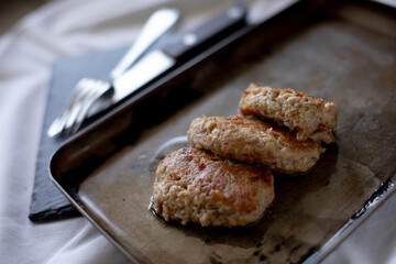 Freshly fried meat patties rest on a slightly worn metal tray, with visible oil drops enhancing their golden appearance. The cutlery peeks from the background on a dark surface, giving a rustic, homel