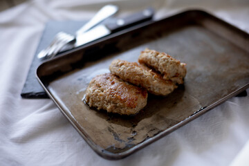 Freshly fried meat patties rest on a slightly worn metal tray, with visible oil drops enhancing their golden appearance. The cutlery peeks from the background on a dark surface, giving a rustic, homel