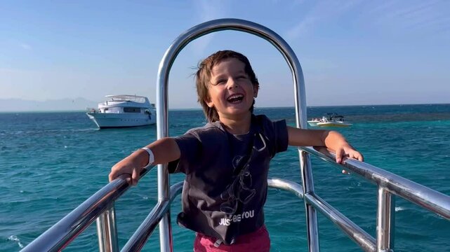 A cute pretty little boy stands at the stern of a yacht sailing on a sunny day against the background of other yachts. The child is happy and laughing, his hair is fluttering in the wind. High quality