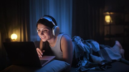 A woman is happily enjoying late night streaming entertainment on her laptop while wearing headphones in a cozy setting - Powered by Adobe
