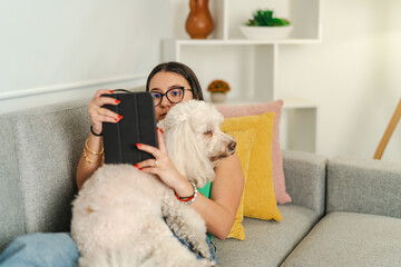 Young woman relaxing on sofa reading e-book with pet poodle