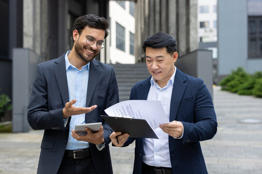 Two business professionals collaborating reviewing documents outdoors. One holds a tablet while they discuss details regarding a clipboard with printed papers. They appear engaged and happy working