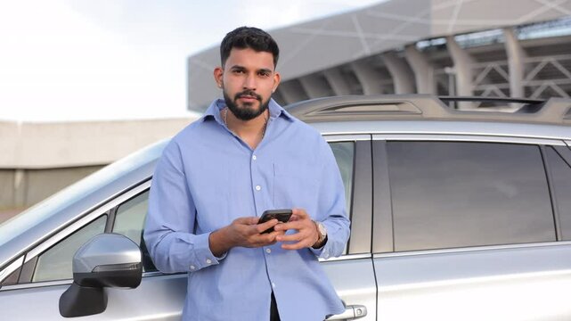 Handsome strong dreamy man using smartphone, while leaning on glossy car outdoors. Portrait of attractive Caucasian driver looking up as camera approaching, waiting near stadium. Concept of lifestyle.