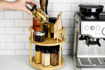 hand is placing or removing a small jar from the top tier. The background features a white tiled wall and a coffee machine.