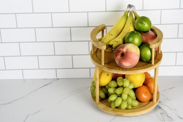 The image shows a two-tier wooden fruit stand on a white marble countertop against a white tiled wall. The top tier holds bananas, limes, and peaches, while the bottom tier contains lemons,