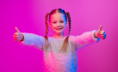 A preteen girl with braided hair smiles brightly while giving two thumbs up against a vibrant neon...