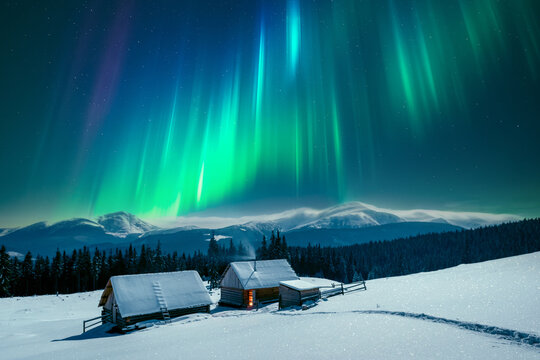 A winter scene with a solitary wooden cabins in snowy mountains under incredible night sky with Northern lights. Aurora borealis. Christmas holiday and winter vacations concept