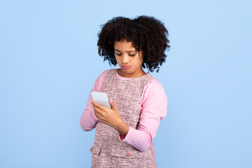 A joyful preteen black girl stands isolated in front of a blue backdrop, engaged with her phone. Her curly hair frames her face, reflecting her happiness and vibrant energy.