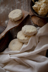 A rustic wooden table with cottage cheese pancakes (syrniki) on a wooden spoon, accompanied by a bowl of fresh cottage cheese. The flour-dusted surface creates a cozy and warm atmosphere