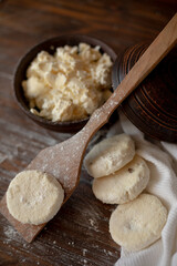 A rustic wooden table with cottage cheese pancakes (syrniki) on a wooden spoon, accompanied by a bowl of fresh cottage cheese. The flour-dusted surface creates a cozy and warm atmosphere