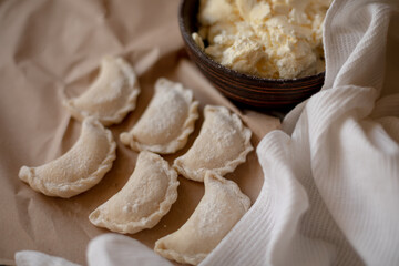 Fresh dumplings (varenyky) filled with cottage cheese, arranged on parchment paper alongside a bowl of cottage cheese and a white kitchen towel. The wooden background and flour-dusted details 