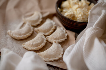 Fresh dumplings (varenyky) filled with cottage cheese, arranged on parchment paper alongside a bowl of cottage cheese and a white kitchen towel. The wooden background and flour-dusted details 