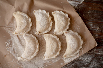 Six handmade dumplings (varenyky) filled with cottage cheese, neatly arranged on parchment paper and dusted with flour. The top-down view and wooden background add a cozy, homemade feel