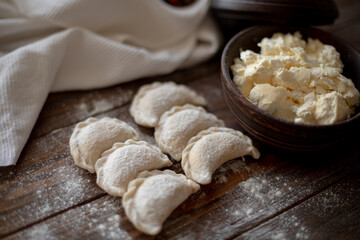 Fresh dumplings (varenyky) filled with cottage cheese, arranged on parchment paper alongside a bowl of cottage cheese and a white kitchen towel. The wooden background and flour-dusted details 