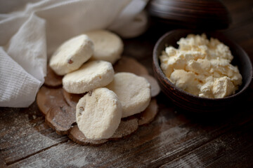 Raw cottage cheese pancakes (syrniki) with raisins, dusted with flour, arranged on a rustic wooden board alongside a dark wooden bowl filled with fresh cottage cheese. A cozy kitchen atmosphere 