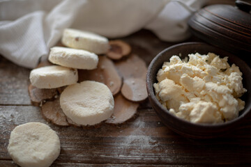 Raw cottage cheese pancakes (syrniki) with raisins, dusted with flour, arranged on a rustic wooden board alongside a dark wooden bowl filled with fresh cottage cheese. A cozy kitchen atmosphere 