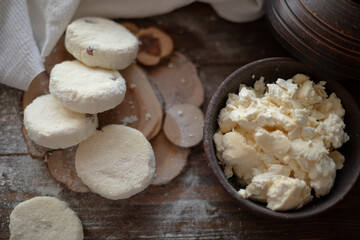 Raw cottage cheese pancakes (syrniki) with raisins, dusted with flour, arranged on a rustic wooden board alongside a dark wooden bowl filled with fresh cottage cheese. A cozy kitchen atmosphere 