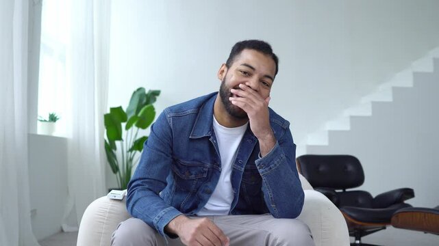 modest african american man is shy and laughs from joke at home, embarrassed guy sits on chair and laughs from compliment