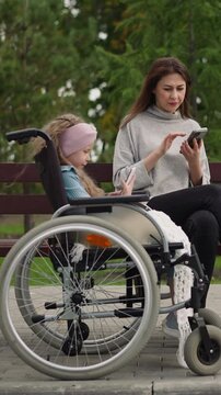 Woman checks email while little girl in wheelchair watching videos on smartphone. Focused mother and daughter use gadgets after walk in city park