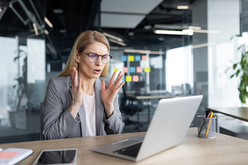 A mature businesswoman with glasses sits at a desk in a modern office, facing frustration during an online meeting. Her gestures communicate discontent as she interacts via laptop.