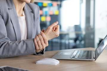 Mature businesswoman seated at an office desk holds her wrist in discomfort, indicating potential repetitive strain injury likely caused by prolonged computer use. T