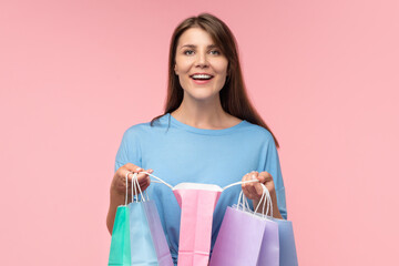 Studio portrait of cute happy smiling casually dressed girl posing over pastel pink background holding a bunch colorful shopping bags in hands and checking her purchase with excited face expression