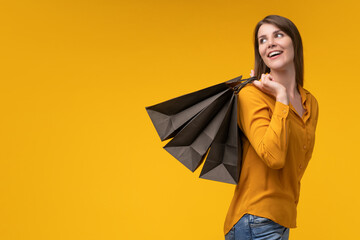 Studio shot of excited young woman customer posing over yellow background holding a bunch of shopping bags, smiling and looking to the side, with the copy space on the left