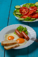 Salad, cucumbers and tomatoes in olive oil and fried eggs with sausages in a plate on a blue wooden table