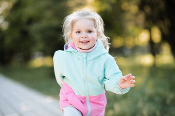 Smiling kid girl 4-5 year old wearing sport jacket running outdoor in park. Looking at camera. Childhood. Springtime.