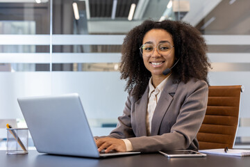 Portrait of a young smiling African American woman in a suit sitting in the office at a desk, using a laptop and looking at the camera