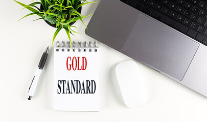 Gold standard written on a notepad with a laptop, pen, mouse, and green plant on a white desk