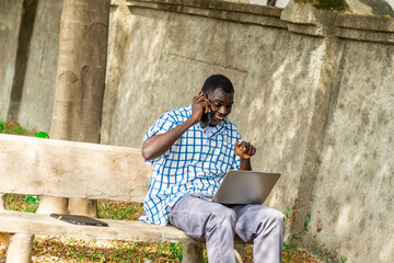 Fototapeta premium Young cheerful black man using mobile phone and laptop computer. Modern lifestyle