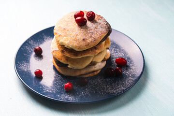 Cottage cheese pancakes with cranberries are stacked on a blue plate on a light blue table, sprinkled with powdered sugar