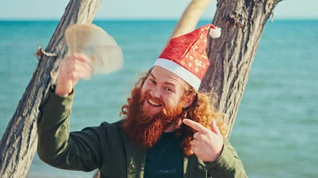  A cheerful bearded man in a Santa hat playfully shaking dollar bills and bragging about his wealth while outdoors.