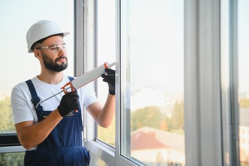 Young man, dressed in a uniform, doing repair in the house. Handyman is applying silicone from a...