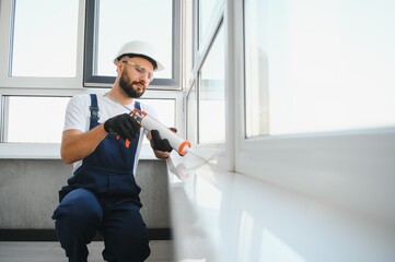 Young man, dressed in a uniform, doing repair in the house. Handyman is applying silicone from a...