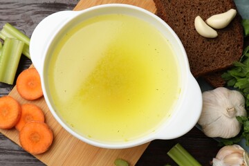 Tasty chicken bouillon in bowl with bread and ingredients on wooden table, flat lay