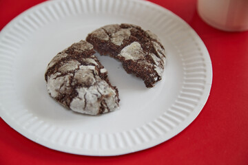 A broken chocolate cookie with cracks lying on a white paper plate against a red background, closeup