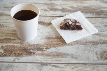 A paper napkin with one half of a chocolate cookie with cracks and a glass of coffee next to it on a wooden table, close-up, half portion, hearty breakfast, limiting sweet and pastry consumption.